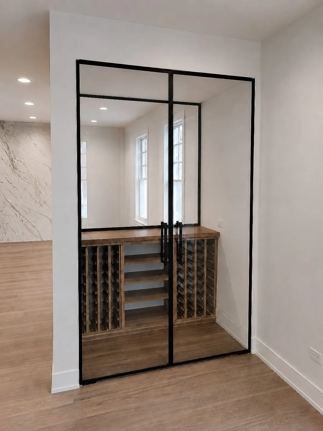 Tall black-framed glass doors with a transom around a wine room with a wood counter and bottle storage.